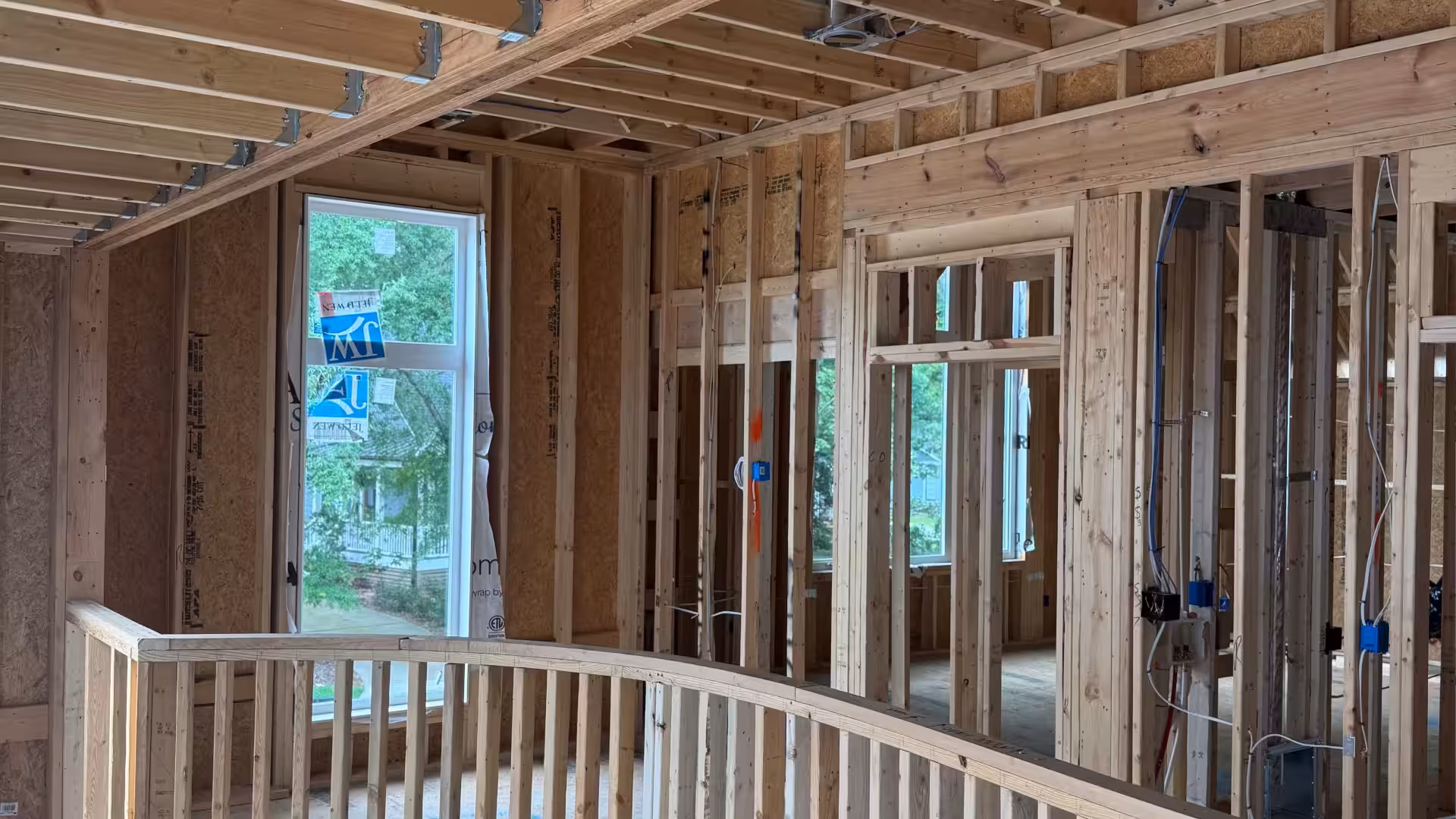 Wood framing of a house under construction with exposed studs, beams, and wiring visible; large windows face an outdoor view with trees.