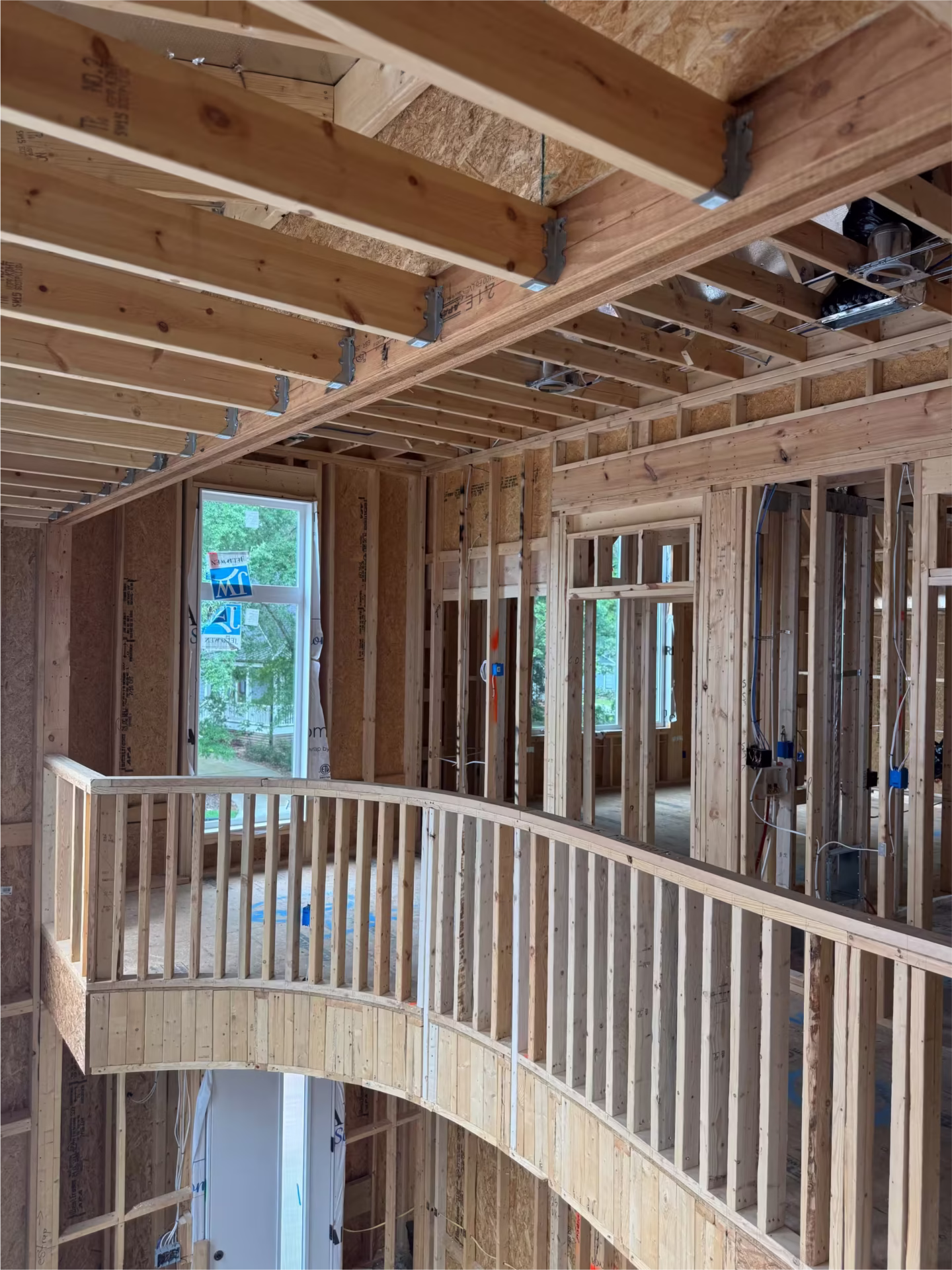 Second floor of a house under construction, showing exposed wooden framing, ceiling beams, an unfinished curved railing, and large windows.