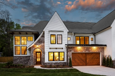Two-story modern house with white siding, brick accents, large windows, and a wooden double garage door, illuminated by exterior lights at dusk.
