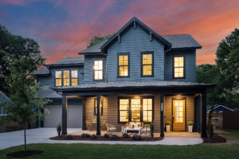 Two-story house with dark blue siding, lit windows, a covered front porch with seating, and a driveway, set against a colorful sunset sky.