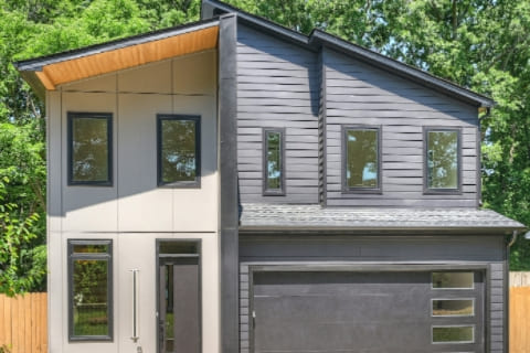 Two-story modern house with asymmetrical roof, dark panel siding, multiple rectangular windows, and a double garage door, set against a background of green trees and a wooden fence.